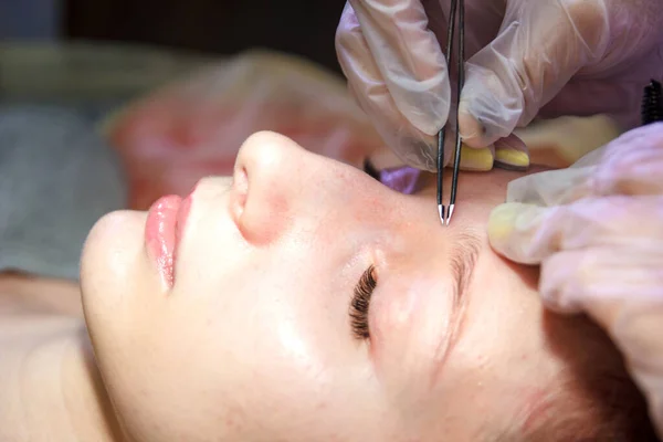Un esteticista en un salón de belleza arranca las cejas en la cara de un cliente con pinzas — Foto de Stock