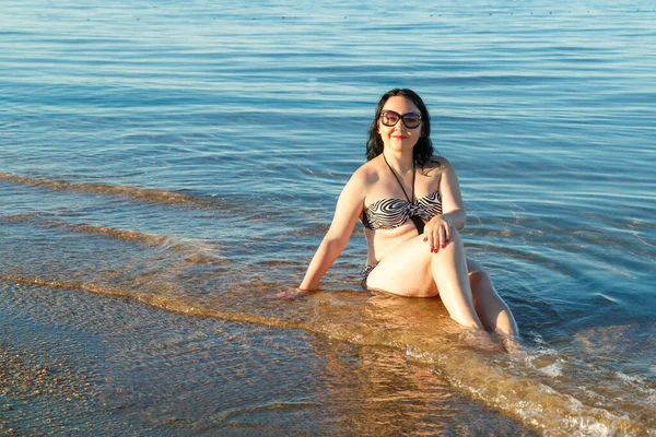 Een brunette vrouw in een badpak en bril zit op de kust. — Stockfoto