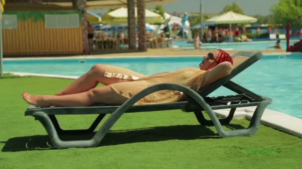 En ung kvinna i en badrock sunbathes på en solstol nära poolen. — Stockvideo