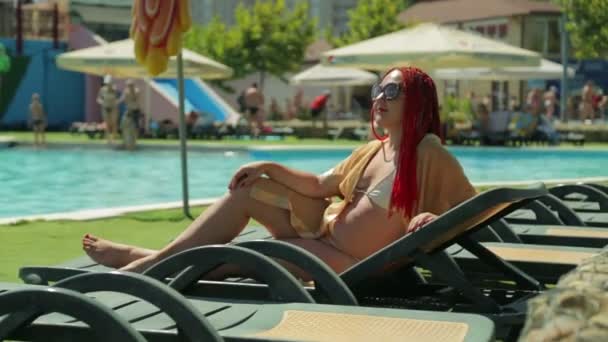Eine Frau mit roten Haaren sonnt sich auf einer Liege am Pool — Stockvideo