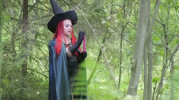 Svůdný mladý čaroděj s červenými vlasy a špičatým kloboukem a s černým pláštěm s svíčkami v lese — Stock video