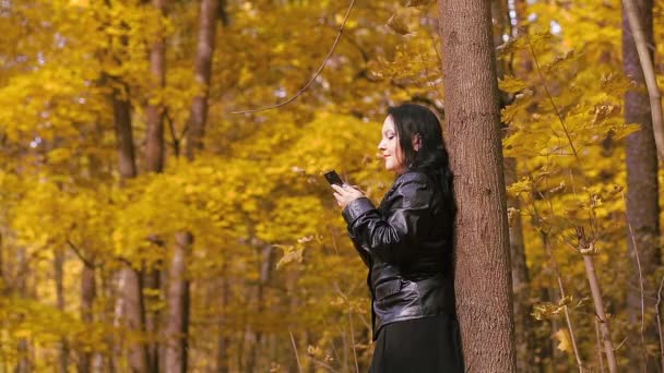 Mladá brunetka v podzimním parku stojí u stromu a píše do telefonu — Stock video