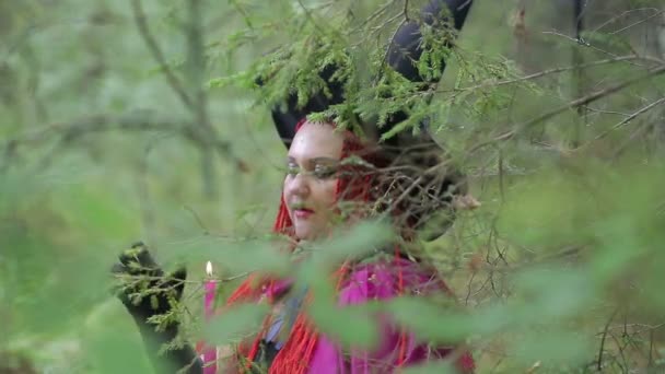 Молодая ведьма с рыжими волосами и черной шляпой и плащом вызывает в лесу свечи — стоковое видео