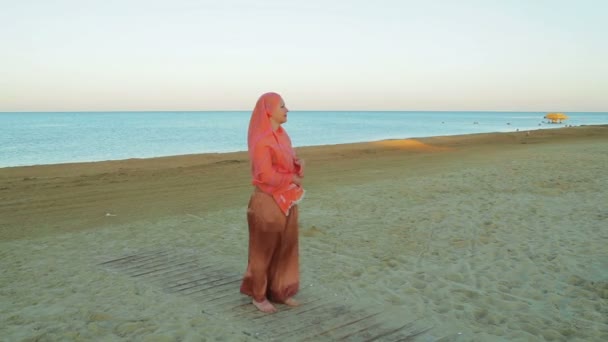 A young Muslim woman in an orange scarf stands on the shore against the background of the sea. — Stock Video