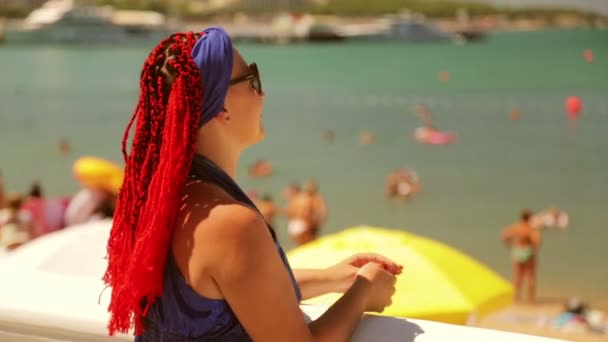 Μια νεαρή γυναίκα με κασκόλ και γυαλιά στέκεται στον παραλιακό δρόμο και κοιτάζει τη θάλασσα και την παραλία. — Αρχείο Βίντεο