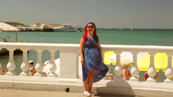 Жінка в блакитній сукні стоїть на набережній і вказує на море — стокове відео