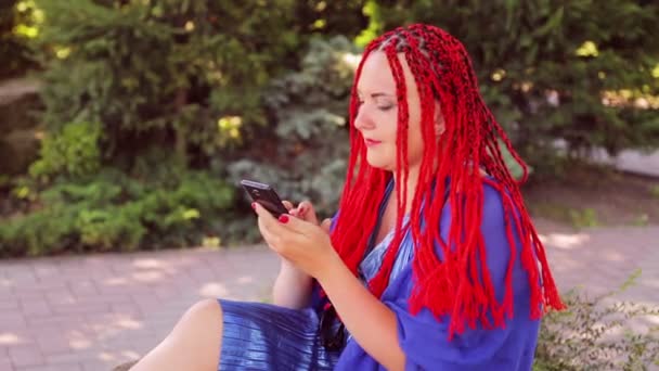 Een jonge vrouw zit op het gras in het park en communiceert in de boodschapper in de smartphone — Stockvideo