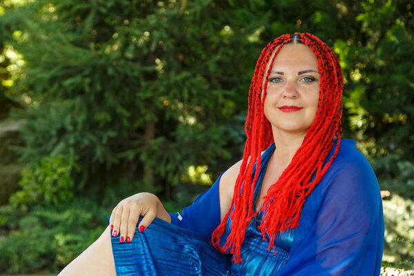 A woman with African pigtails in a blue dress is sitting on the grass in a park.
