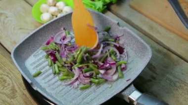 Bir kadın eli, soğanla yeşil fasulyeyi ahşap bir spatulayla karıştırıyor.