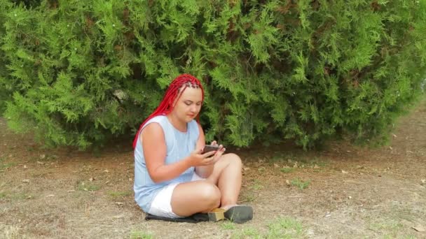 Una joven con coletas rojas está sentada en un parque en la hierba mirando su teléfono — Vídeos de Stock