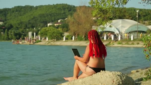 En kvinna med rött hår är blogga på stranden mot bakgrund av bergen — Stockvideo