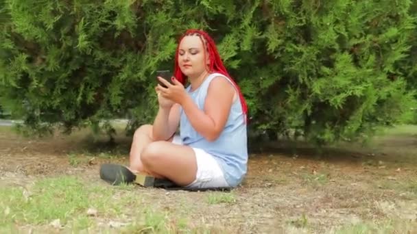 Una joven con coletas rojas está sentada en un parque en la hierba mirando su teléfono. Cámara móvil — Vídeos de Stock