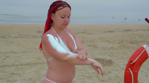 Una giovane donna dai capelli rossi in costume da bagno bianco sta applicando la protezione solare sul corpo sulla riva del mare . — Video Stock