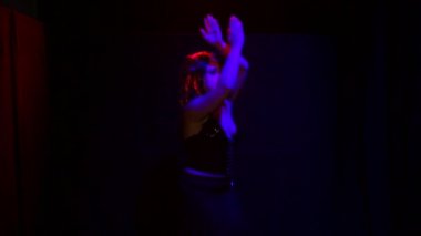 Uğursuz bir gülümsemesi olan şeytan kostümlü bir kadın siyah arka planda kırmızı ışıkta dans ediyor.