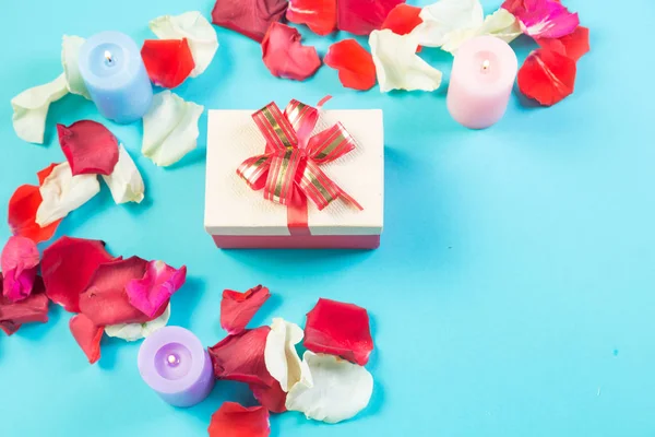 Mavi arka planda gül yaprakları ve kırmızı fiyonklu bir hediye kutusu..