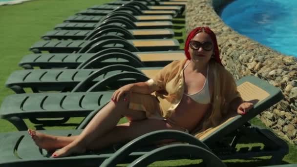 Egy vörös hajú nő napozik egy napozóágyon a medence mellett. — Stock videók