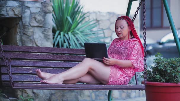 Mujer joven con el pelo rojo en un traje de baño en la playa en una silla de cubierta trabajando con un ordenador portátil — Vídeo de stock