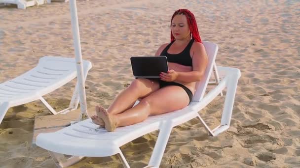 Jeune femme aux cheveux roux en maillot de bain sur la plage sur une chaise longue travaillant avec un ordinateur portable — Video