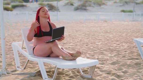 Young woman with red hair in a swimsuit on the beach on a deck chair working with a laptop. — Stock Video