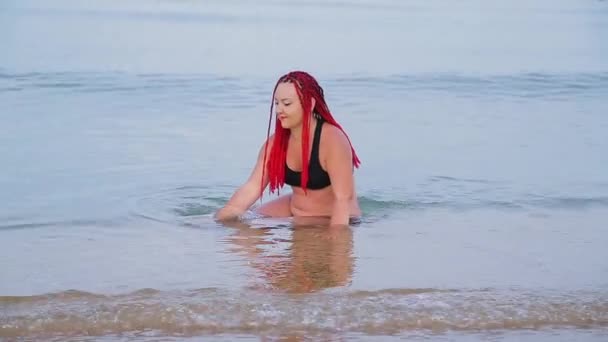 Женщина в черном купальнике с красными косичками сидит в море и наслаждается — стоковое видео