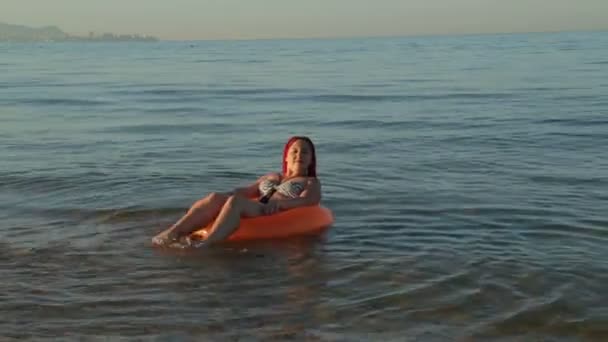 A woman with red hair in a bathing suit on the sea waves in a swimming circle sunbathes. — Stockvideo