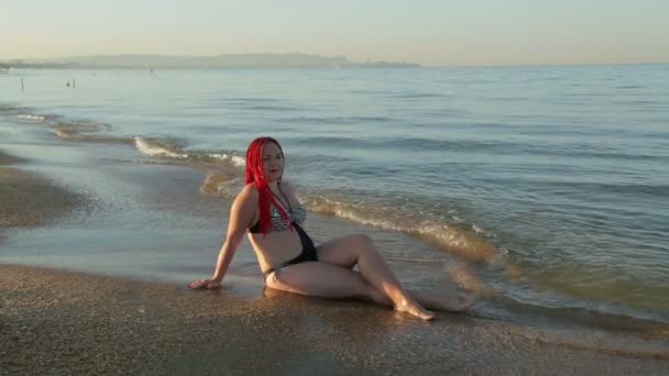 Egy nő a fürdőruhát ül a tengerparton, amelyen hullámok futnak — Stock videók