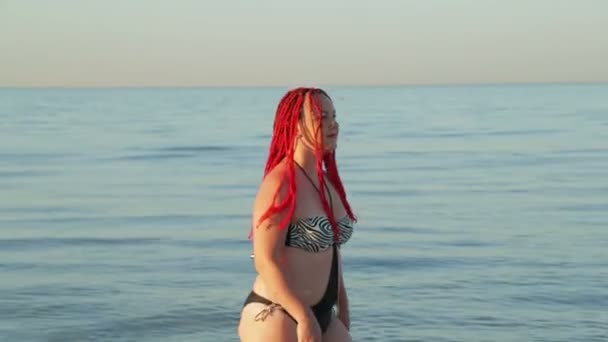 빨간 머리를 한 수영복을 입은 여성이 해안을 따라 걷고 있습니다. — 비디오
