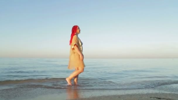 Una donna in costume da bagno e un accappatoio giallo in mare cammina lungo la spiaggia — Video Stock