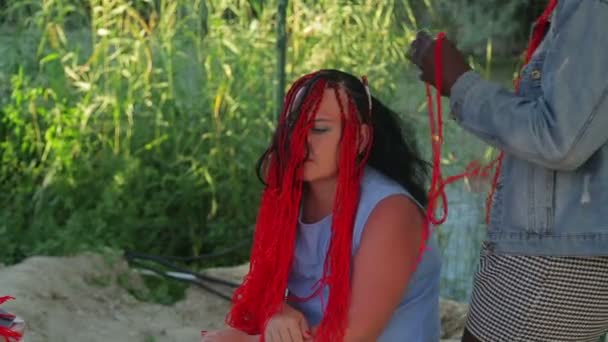 Un coiffeur à la peau foncée tresse une femme avec des tresses avec des fils rouges — Video