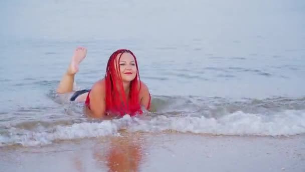 A woman in a black swimsuit with red braids lies on the seashore in sea water — 비디오