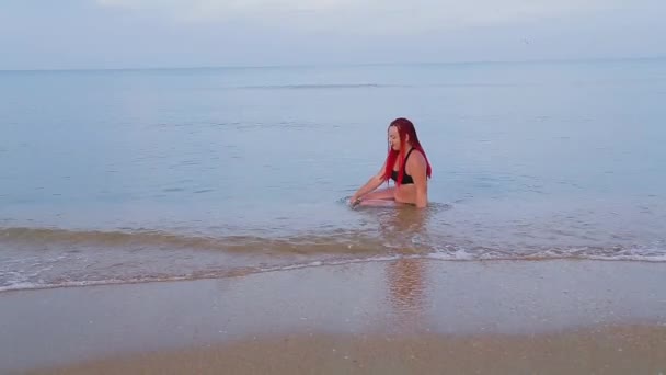 A woman in a black swimsuit with red braids sits in the sea and enjoys — 비디오