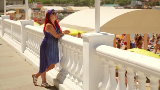 Una mujer con un vestido azul en el paseo marítimo se para y mira a la playa y al mar — Vídeo de stock