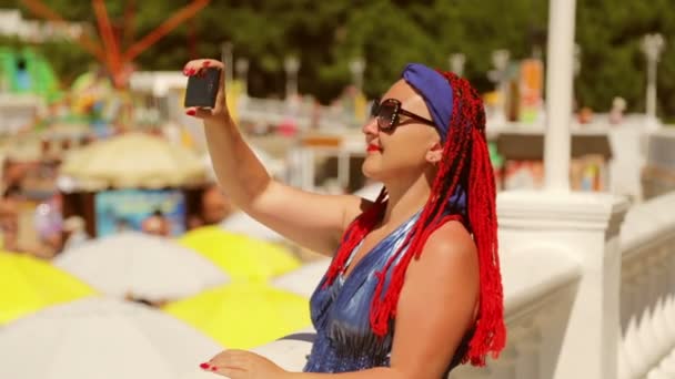 Een jonge vrouw met Afrikaanse vlechten en glazen staat op de promenade en neemt een selfie — Stockvideo