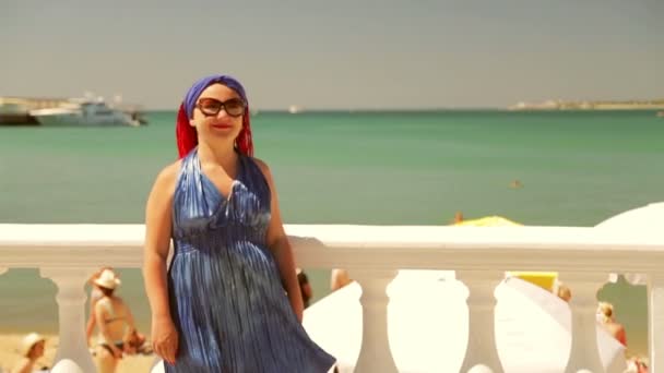 Una mujer con un vestido azul se para en el paseo marítimo y señala el mar — Vídeo de stock