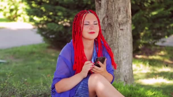 Une jeune femme s'assoit sur l'herbe dans le parc et communique dans le messager dans le smartphone . — Video