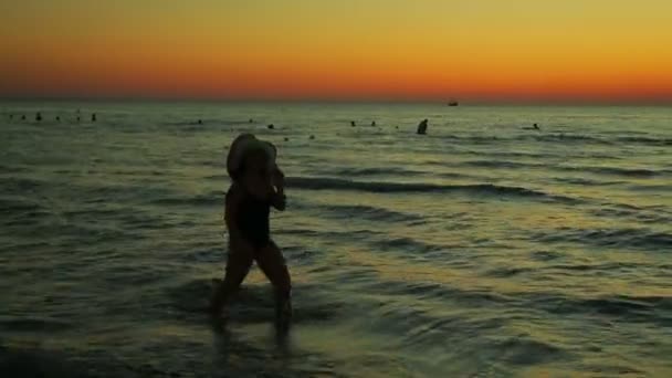 Una donna con un cappello al tramonto cammina lungo la riva del mare. Silhouette — Video Stock