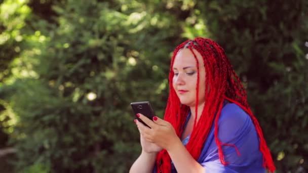 Una mujer joven se sienta en el césped del parque y se comunica con el mensajero en el teléfono inteligente — Vídeo de stock