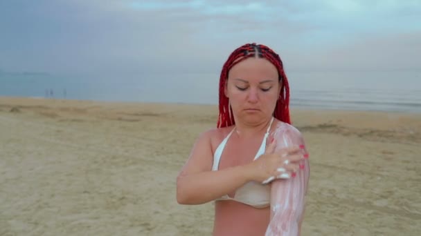 Een jonge roodharige vrouw in een wit badpak brengt zonnebrandcrème aan op het lichaam aan de kust. — Stockvideo