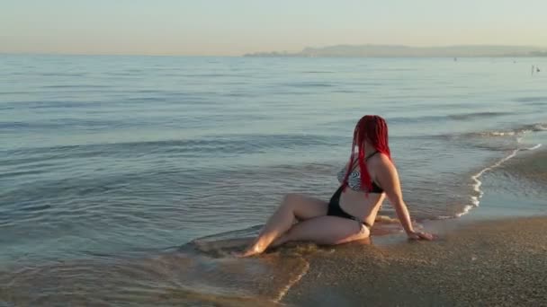 A woman in a swimsuit sits on the seashore on which waves are running — Stock Video