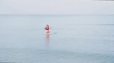 Kırmızı örgülü siyah mayo giymiş bir kadın denizden çıkar.
