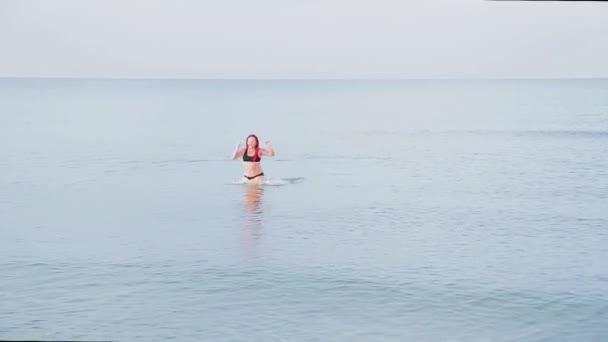 A woman in a black swimsuit with red braids comes out of the sea ashore — Stock Video