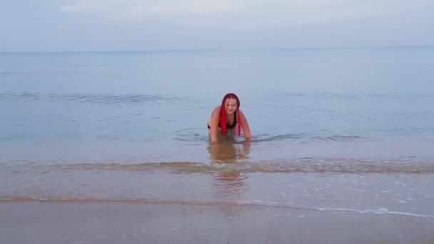 En judisk kvinna i svart baddräkt med röda flätor sitter i havet och faller från mötande vågor — Stockvideo