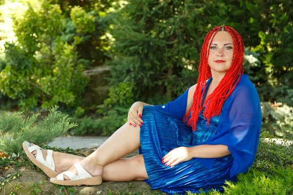 Eine Frau mit afrikanischen Zöpfen im blauen Kleid sitzt auf dem Gras in einem Park. — Stockfoto