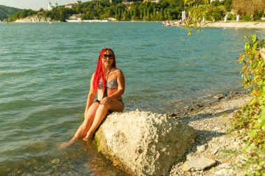 Mayo içinde Afrikalı pigtails ile genç bir kadın bir dağ gölü kıyısında bir taş üzerinde oturur.