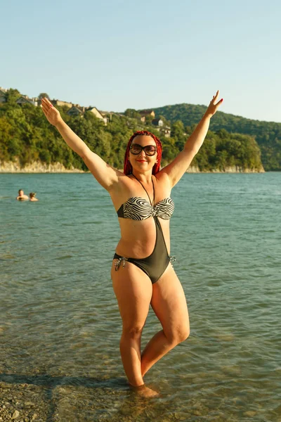 Una hermosa mujer con coletas africanas en traje de baño está de pie sobre una piedra junto al lago sobre un fondo de montañas . — Foto de Stock