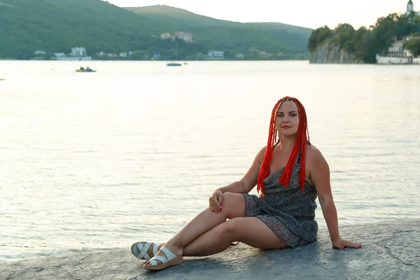 Vacker kvinna med rött hår sitter vid sjön mot bakgrund av berg vid solnedgången. — Stockfoto