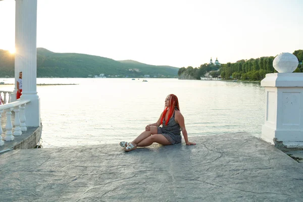 Hermosa mujer con el pelo rojo se sienta junto al lago en el fondo de las montañas al atardecer . — Foto de Stock