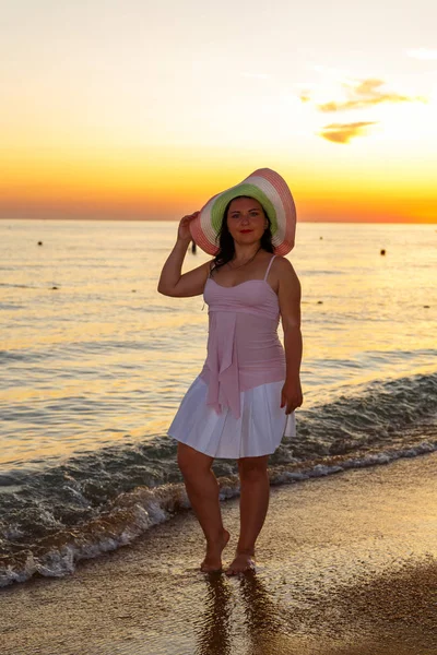 Una joven con un vestido blanco y un sombrero en la orilla del mar al atardecer . — Foto de Stock