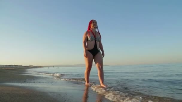 Een vrouw in een badpak met rood haar wandelingen langs de kust — Stockvideo