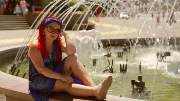 Triste mujer en un vestido azul con coletas sentado en la fuente — Vídeos de Stock